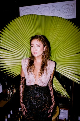 Ashley Park in a white-black lace dress and black lace gloves posing in front of a large plant