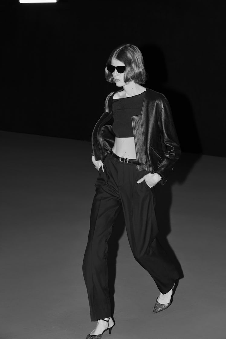 A female model walking while wearing a black top, black pants, a black leather jacket, and black sun...