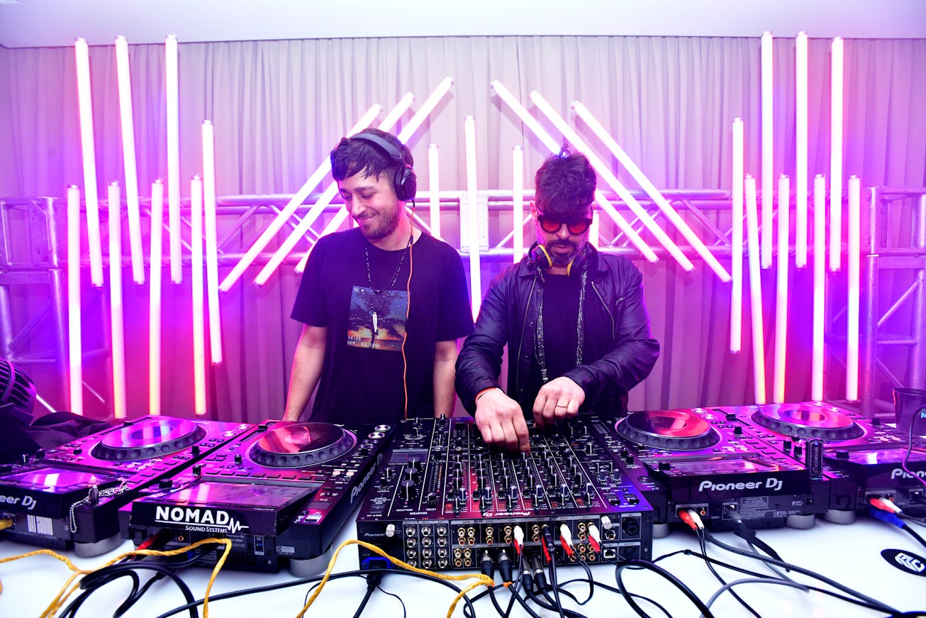   Two male DJs wearing black and standing next to a mixing board, inside the NYLON house at Art Base...