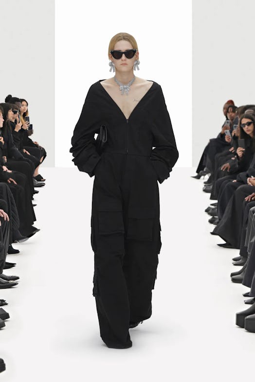 Model walking down a runway while wearing a full black Balenciaga jumpsuit, jewelry, and black glass...