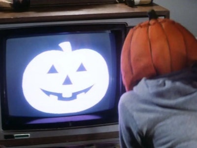 A person with a pumpkin head watching TV with a Halloween pumpkin on TV screen