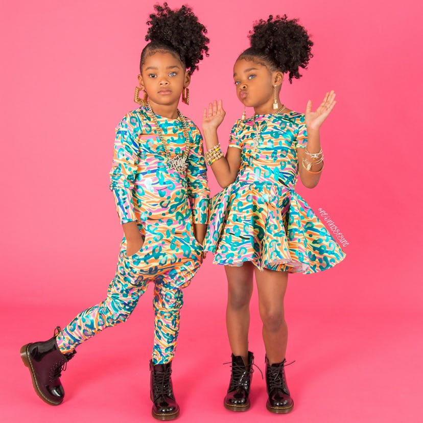 two girls in front of pink backdrop, one wearing jumpsuit the other in a dress both of which are in ...