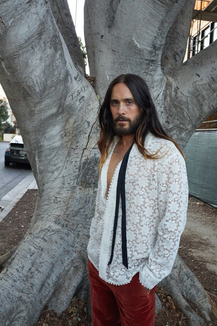 Jared Leto in a white floral shirt and red pants posing in front of a tree