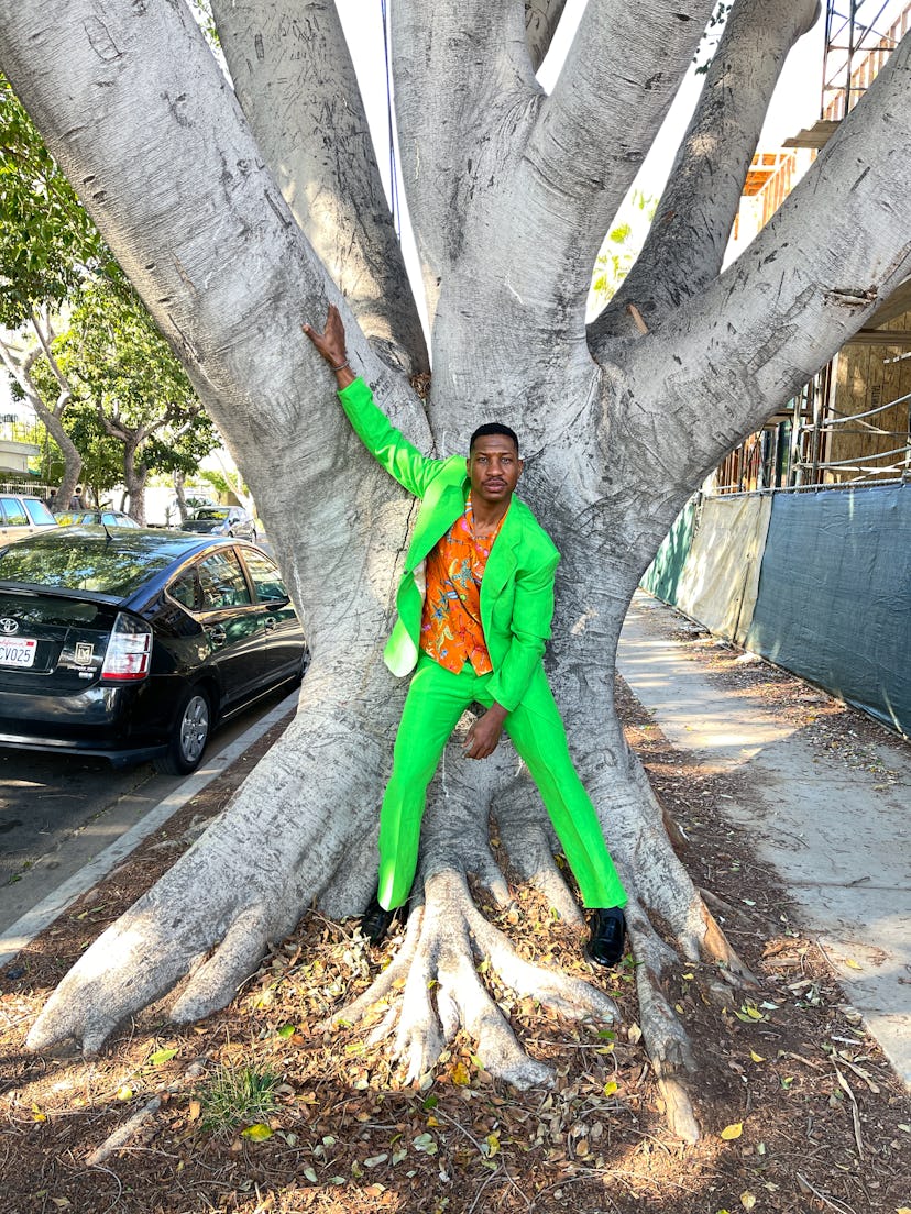 Jonathan Majors in a green Versace suit and orange shirt, leaning against a tree