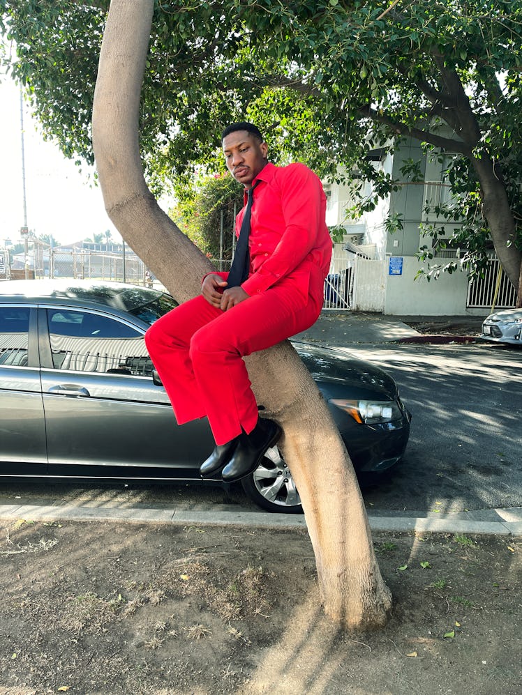 Jonathan Majors in a red button-up, matching pants and a black tie, sitting on a tree branch 