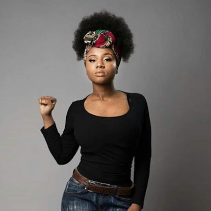 Nupol Kiazolu, wearing a black shirt, jeans, and colorful headband wrap, stands in front of a gray b...