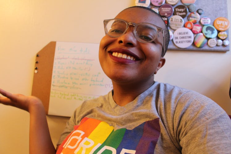 A selfie of Ose Arhegan, wearing a colorful Pride shirt, smiling into a camera in front of a whitebo...