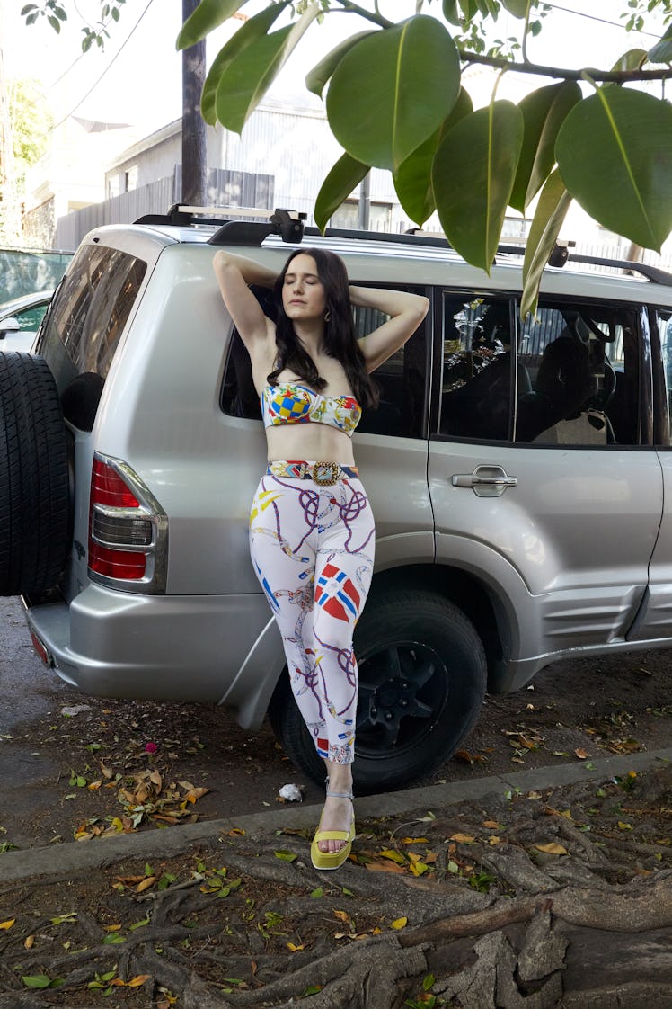 Rachel Brosnahan in a bikini top and leggings with a pattern-print while leaning onto a white vehicl...