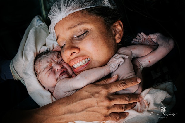 A woman cradles a nude newborn child against her cheek moments after its birth. 