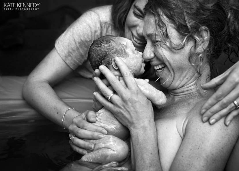 A nude woman smiles as she touches her nose to the nose of a newborn child while another woman smile...