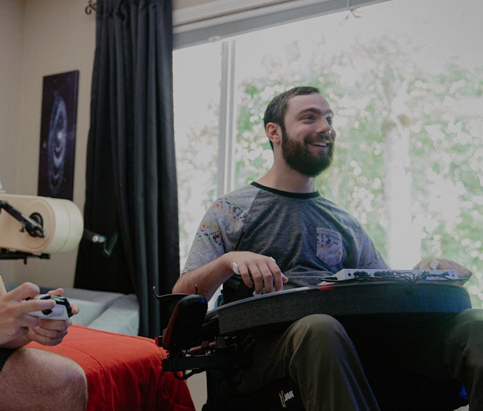 Two gamers are seen sitting together while playing on an Xbox console. One of the gamers is in a whe...
