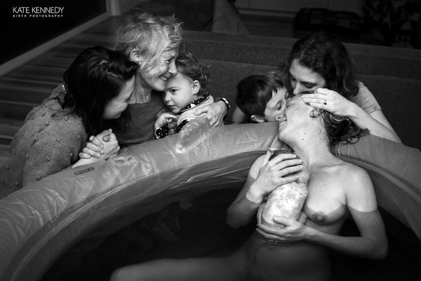 A nude woman cradles a newborn to her chest as she reclines in a pool of water smiling while three w...