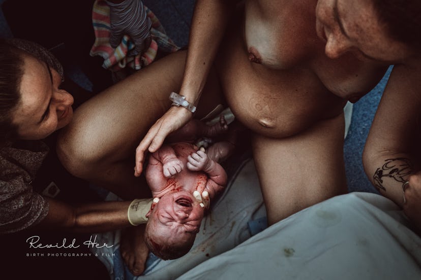 A woman holds a newborn as it is born while the nude laboring mother reaches down to touch the child...