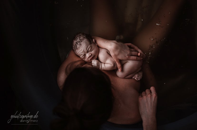 A nude woman with dark hair is pictured from above sitting in water while cradling a nude newborn ch...