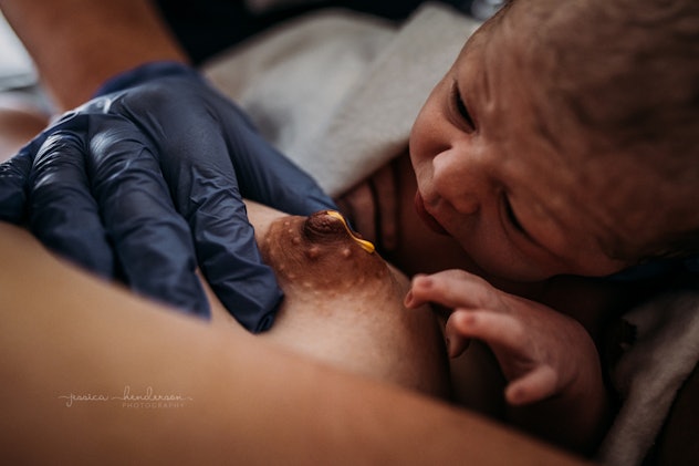 A gloved hand rests on an exposed breast as colostrum drips from the nipple and a newborn looks on.
