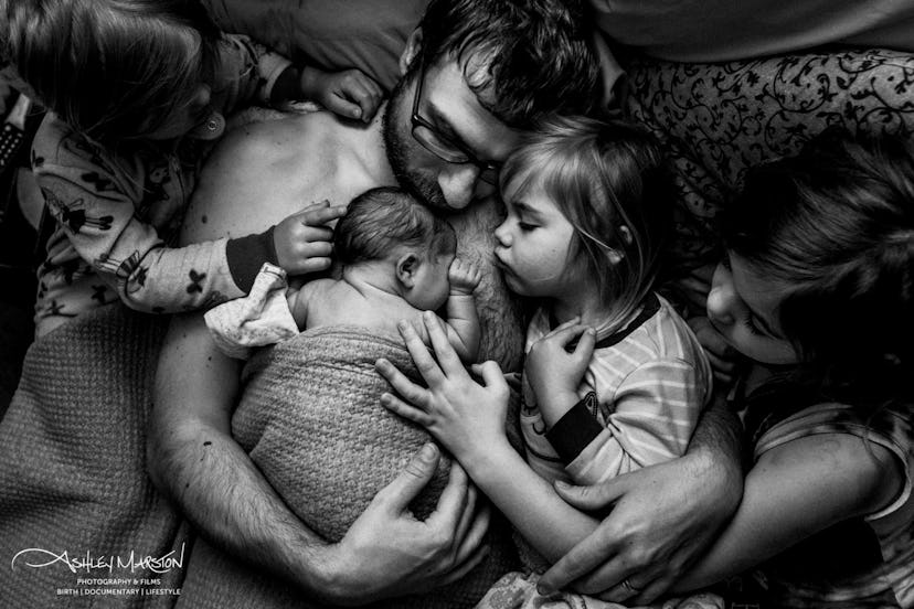 In a black and white photograph, a father cradles a swaddled newborn baby against his bare chest whi...