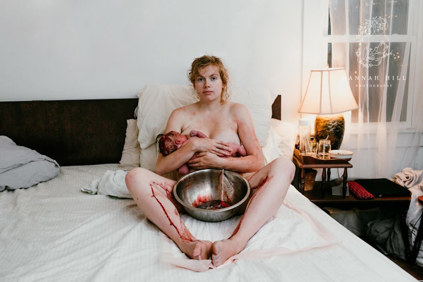 A nude woman reclines on a bed made with white sheets while nursing a newborn baby and cradling a la...