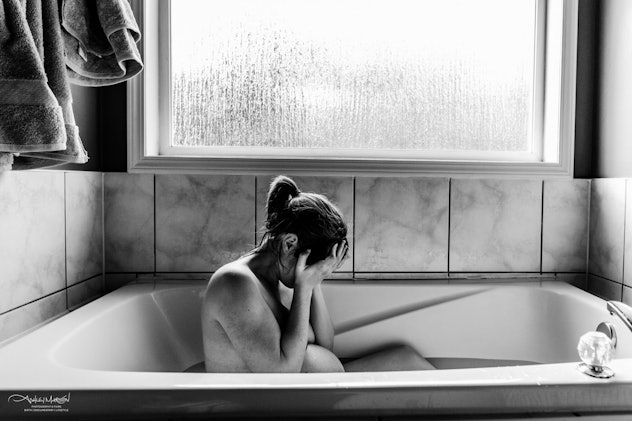 A nude woman cradles her head in her hands while sitting in a half-filled bathtub in this black and ...
