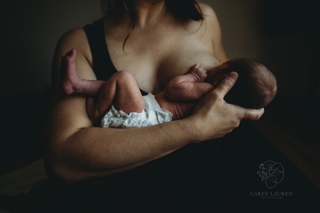 A woman is pictured from the neck down nursing a diaper-clad newborn while wearing a black sleeveles...