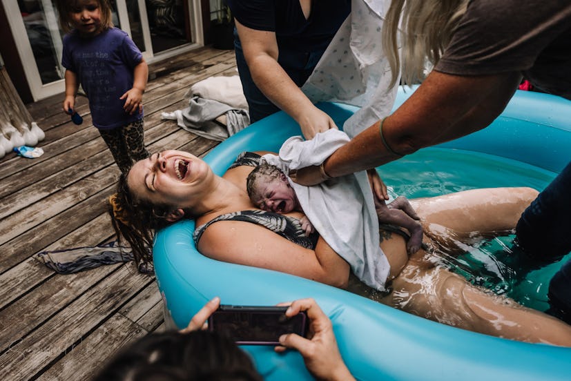 A woman cradles a newborn to her chest as she lies with her head back laughing in a birthing pool as...