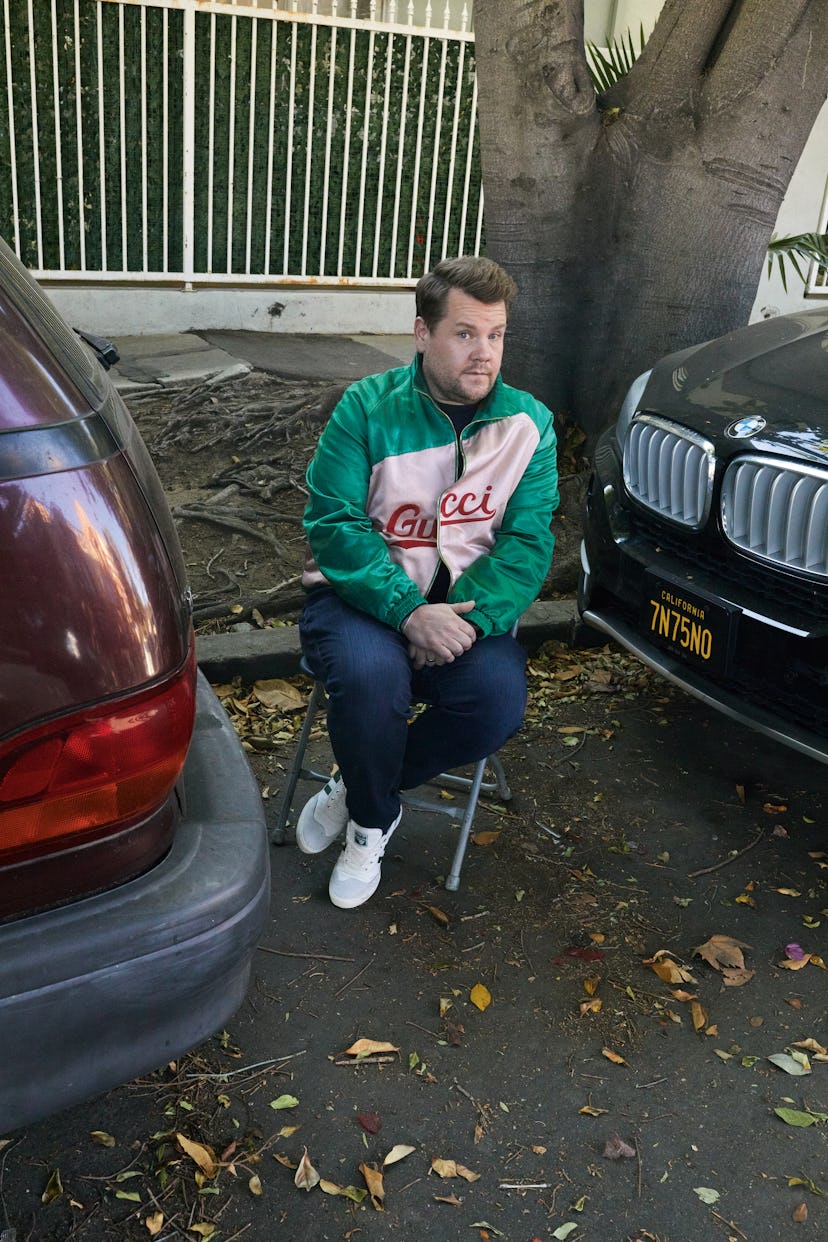 James Corden sitting on a chair between two cars in a green and white Gucci jacket, jeans and white ...