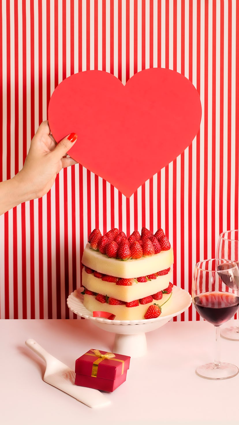 Heart-shaped cake, layered, with strawberries; sitting on white cake stand in front of red and white...