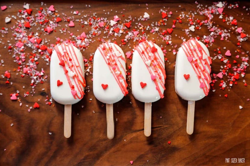Four white cake Popsicles laying on a wood table with red, pink, and white sprinkles and candy heart...