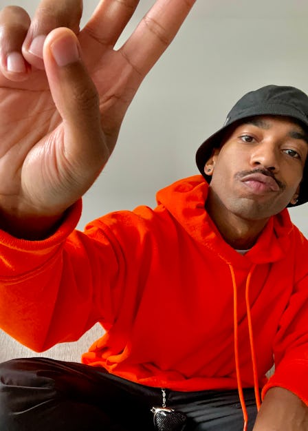 Antoine Gregory in an orange hoodie, black pants and a black bucket hat and his hand showing the pea...