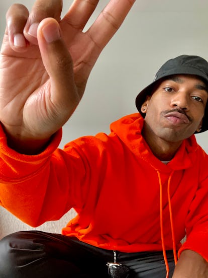 Antoine Gregory in an orange hoodie, black pants and a black bucket hat and his hand showing the pea...