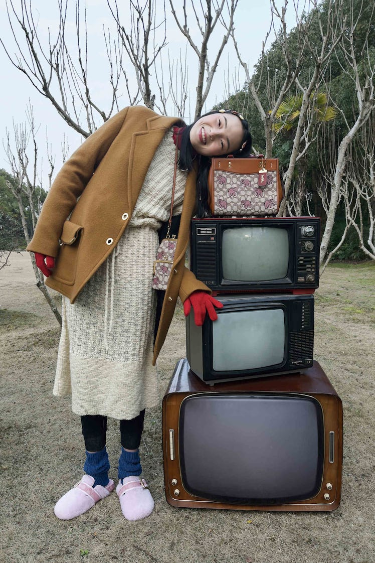 A model leaning against a brown bag stacked on top of old TVs in a beige coat and white knit dress b...