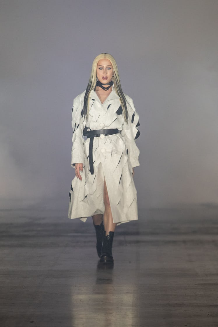A'Whora walking the runway at London Fashion Week in a white and black trench coat with a black belt...