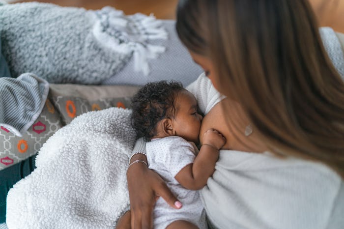 A woman of Indian descent is breastfeeding her baby daughter. The woman is sitting on a couch in her...