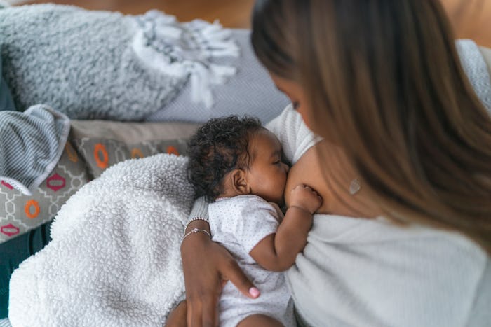 A woman of Indian descent is breastfeeding her baby daughter. The woman is sitting on a couch in her...