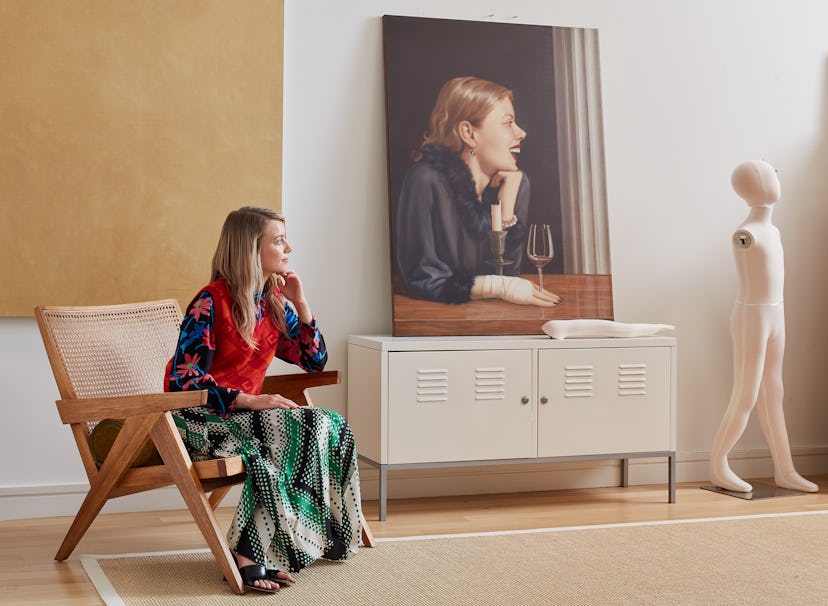 Anna Weyant sitting on a chair in her New York studio mimicking the painting behind her, with her ha...