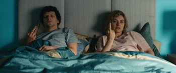 Jesse Eisenberg and Imogen Poots in a blue bed with a grey headboard, their middle fingers raised in disgust.