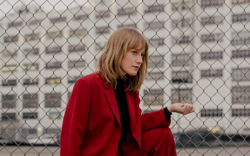 A portrait of The Weather Station's Tamara Lindeman. She's crouched in front of a wire gate in a cri...