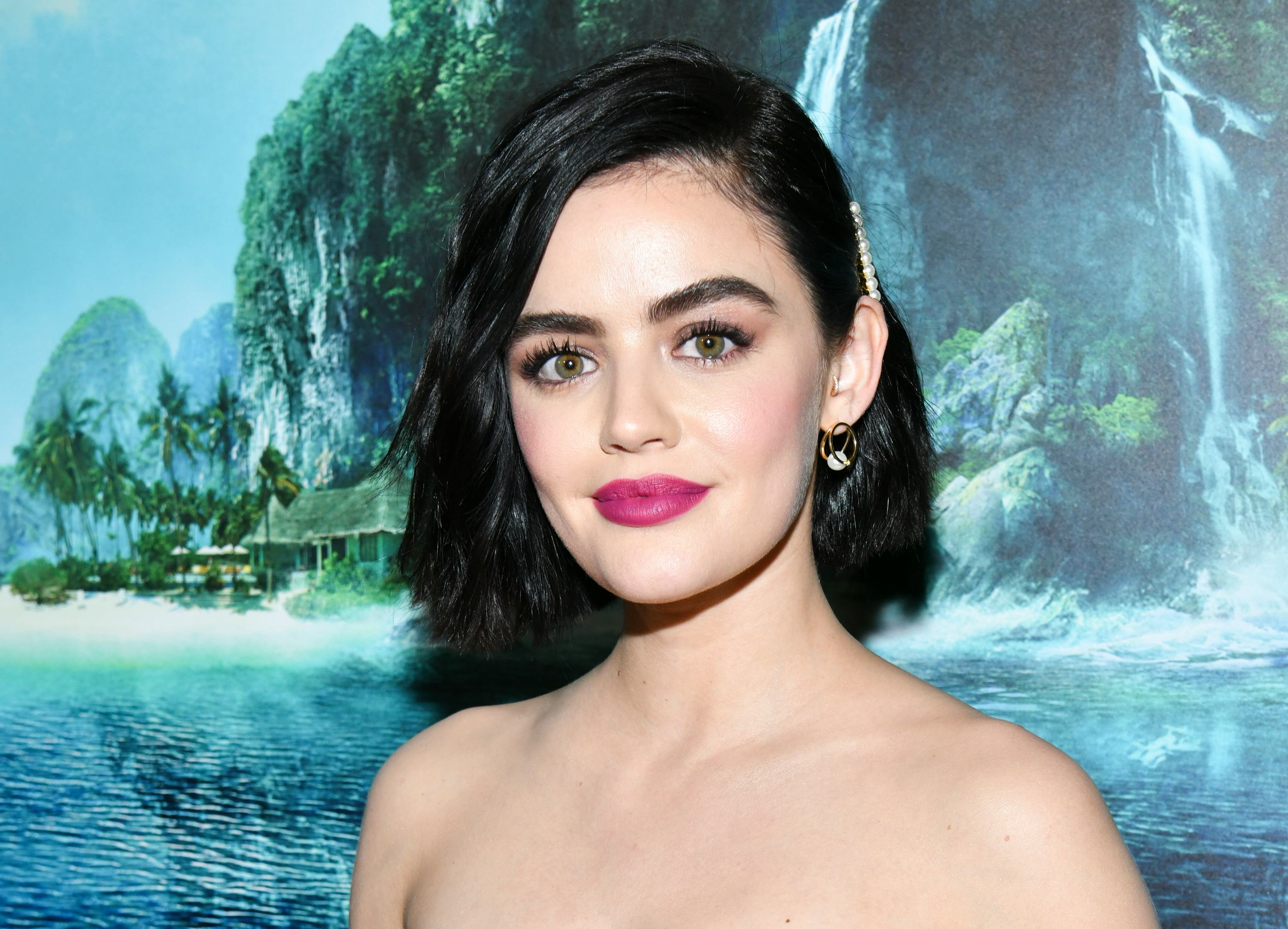 Lucy Hale, wearing pink lipstick, stands against backdrop of fake waterfall