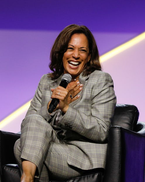 Kamala Harris sitting in a black leather chair laughing while holding a microphone and wearing a gre...