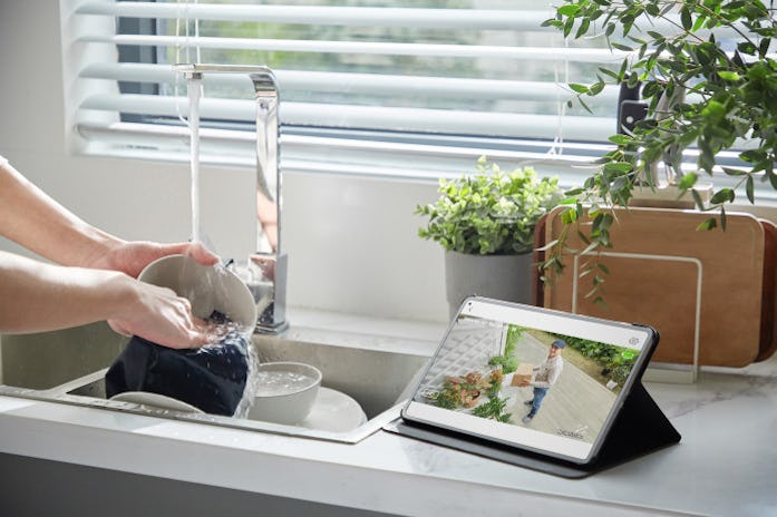 A pair of hands can be seen washing dishes in a kitchen sink. The counter by the sink has a tablet p...