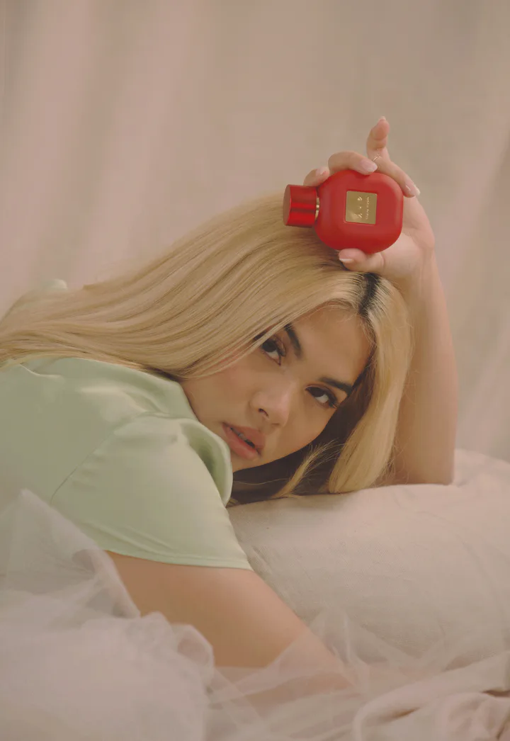 Artist Hayley Kiyoko in mint green blouse holding red bottle of perfume.