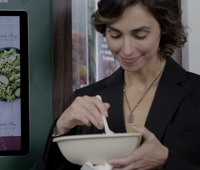 A woman is seen standing in front of a Sally robot by Chowbotics as she eats salad out of a bowl.