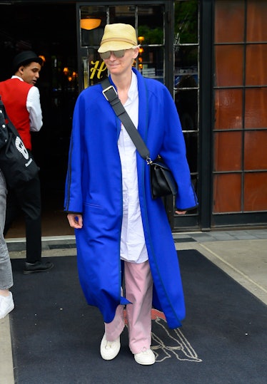 Tilda Swinton leaving a building dressed in an oversized blue coat, pink pants, sunglasses, and a ye...