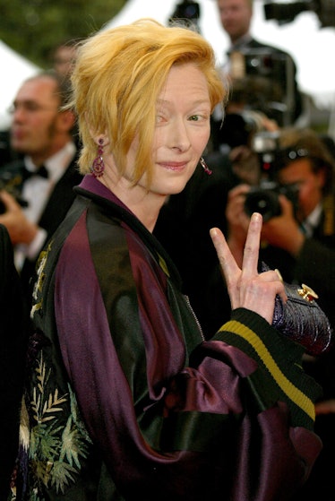 Tilda Swinton showing a peace sign wearing a burgundy and black colored jacket with floral details o...