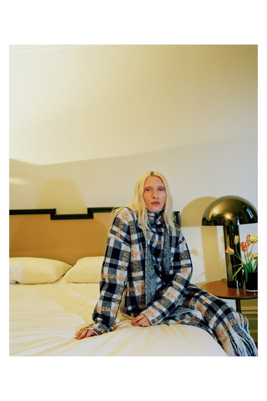 A blonde woman sitting on a bed in a black, blue, yellow, and white plaid shirt