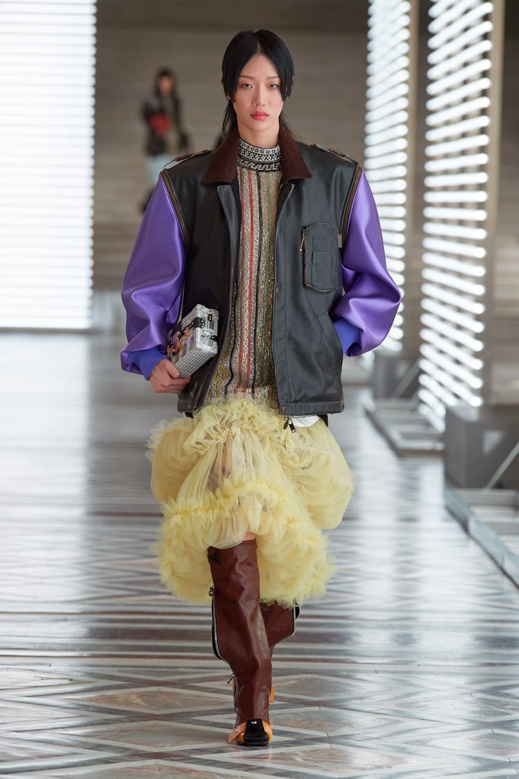 A model walking the runway in a jacket with purple sleeves and a striped dress with tulle on the bot...