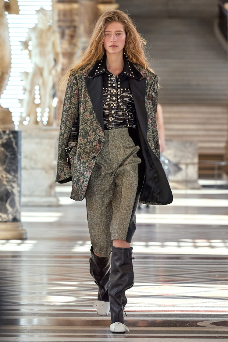 A model walking the runway in a floral jacket, grey pants and black and white boots by Louis Vuitton...