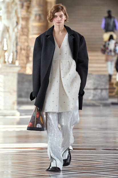 A female model walking in a black coat, white pants, and a white sweater