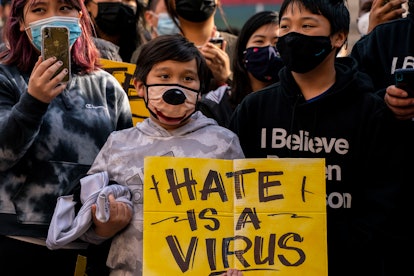 A young Asian child in a face mask holds a sign that says "Hate is a virus." Demonstrators gather in...