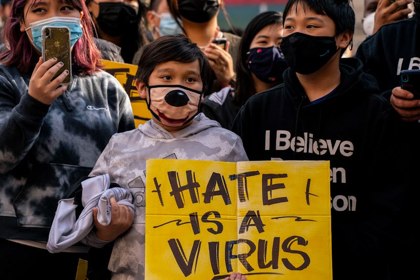 A young child with a face mask holds a sign that says 'HATE IS A VIRUS' in a crowd of people