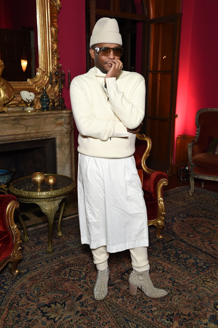 Law Roach standing in a room with his arms crossed while wearing a white skirt and sweater combinati...